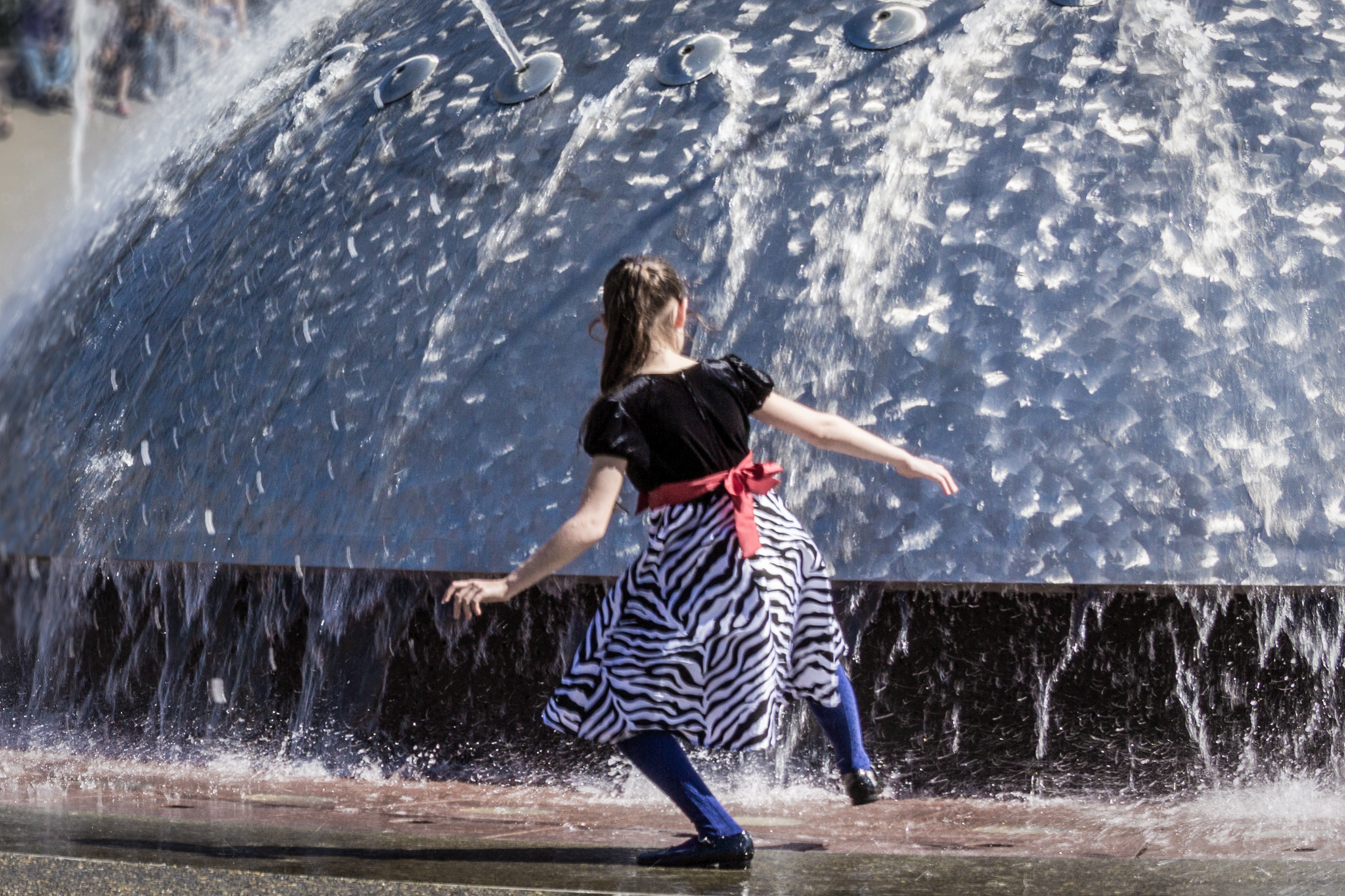 A young girl dressed in nice clothes tips her toes into water of a fountain.