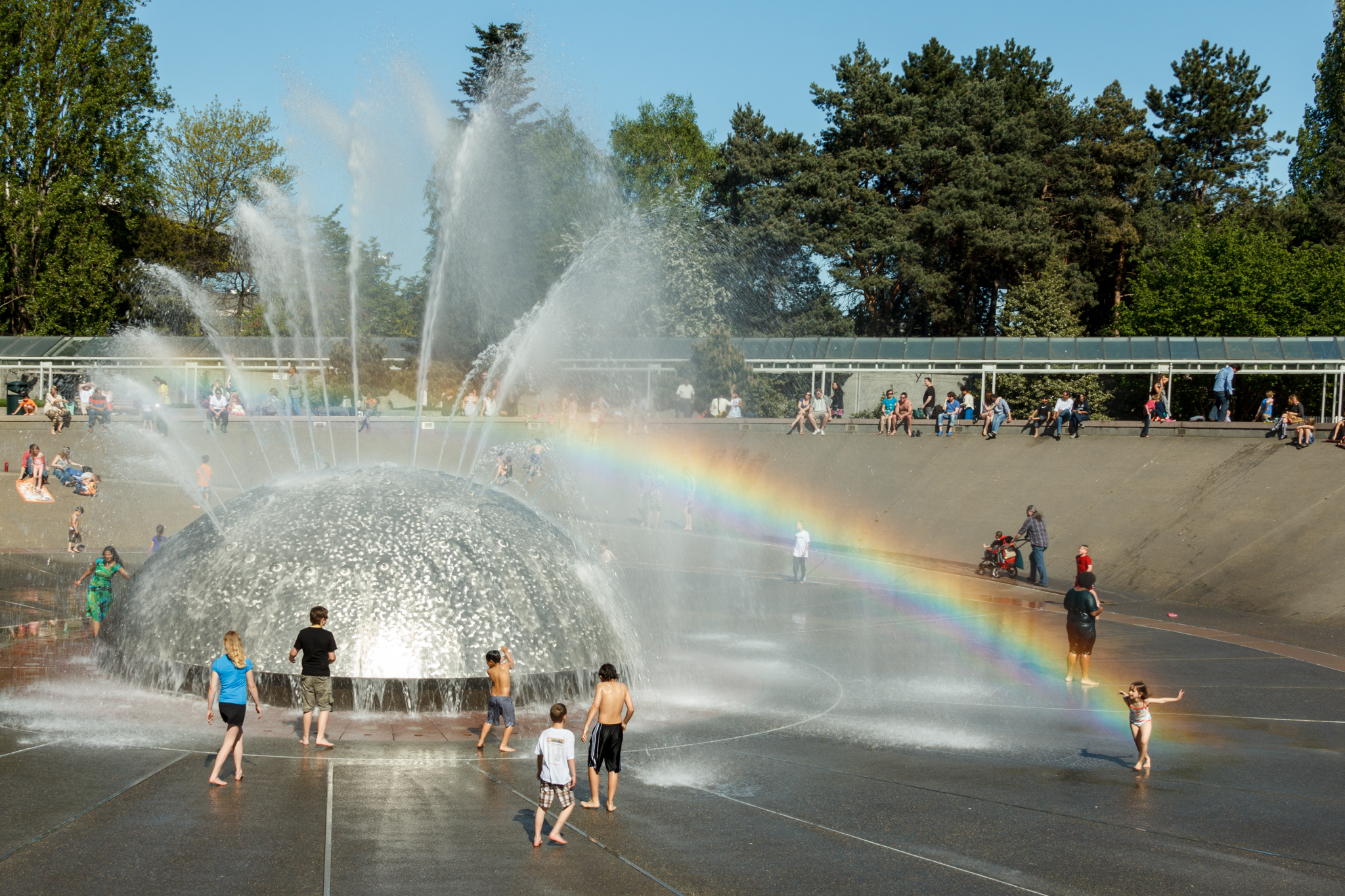 Children dancing in water fountain, one child located at end of rainbow created by fountain spray.