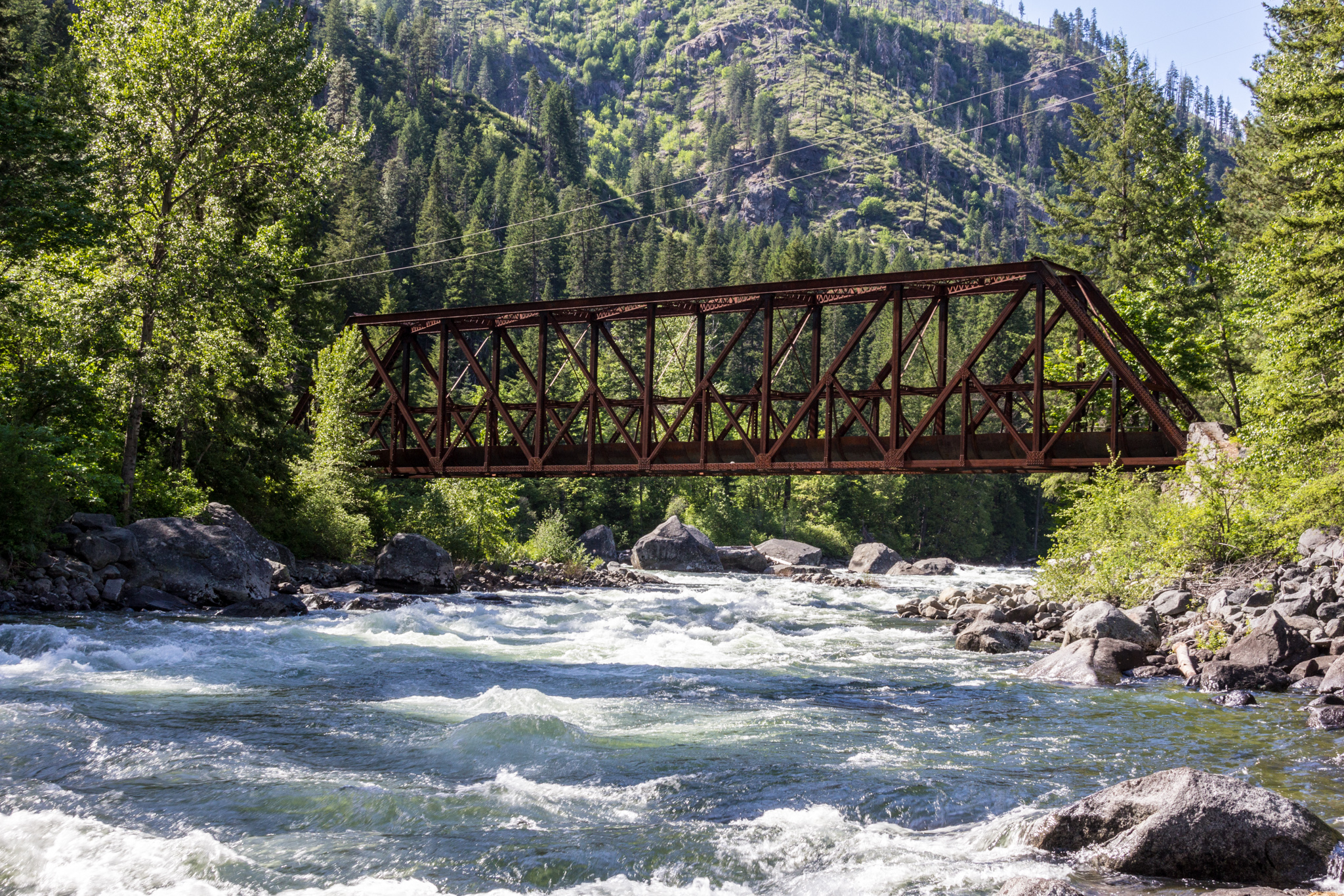 A rusting railroad bridge over river rapids backed by treed hills.